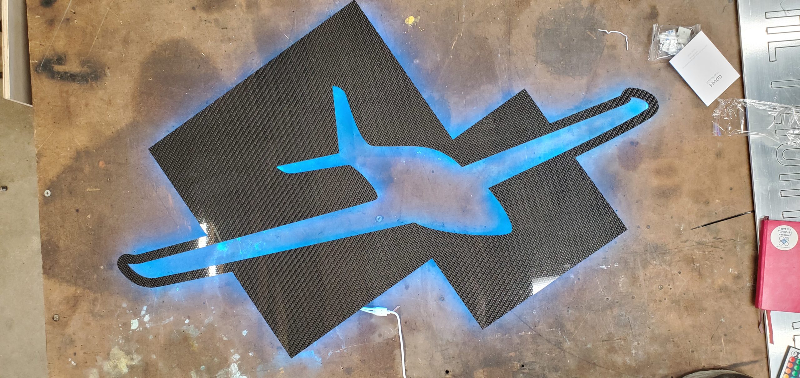 Carbon Fiber logo cutout with a blue glow, sitting on a work table.